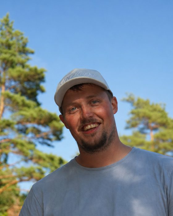 Alexander Lindén i keps och t-shirt, leende, med tallar och blå himmel bakom.